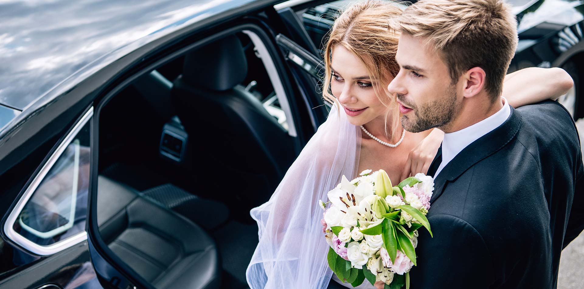 Handsome bridegroom in suit hugging attractive and blonde bride with bouquet